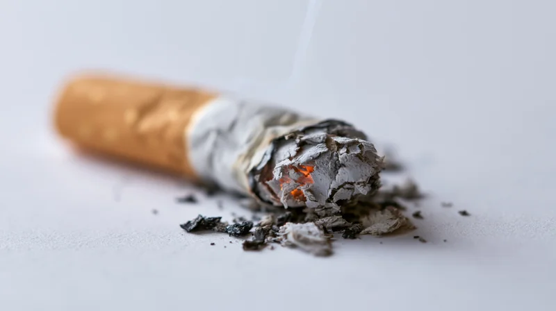 Lit cigarette butt with glowing ember and ash on a plain light grey surface