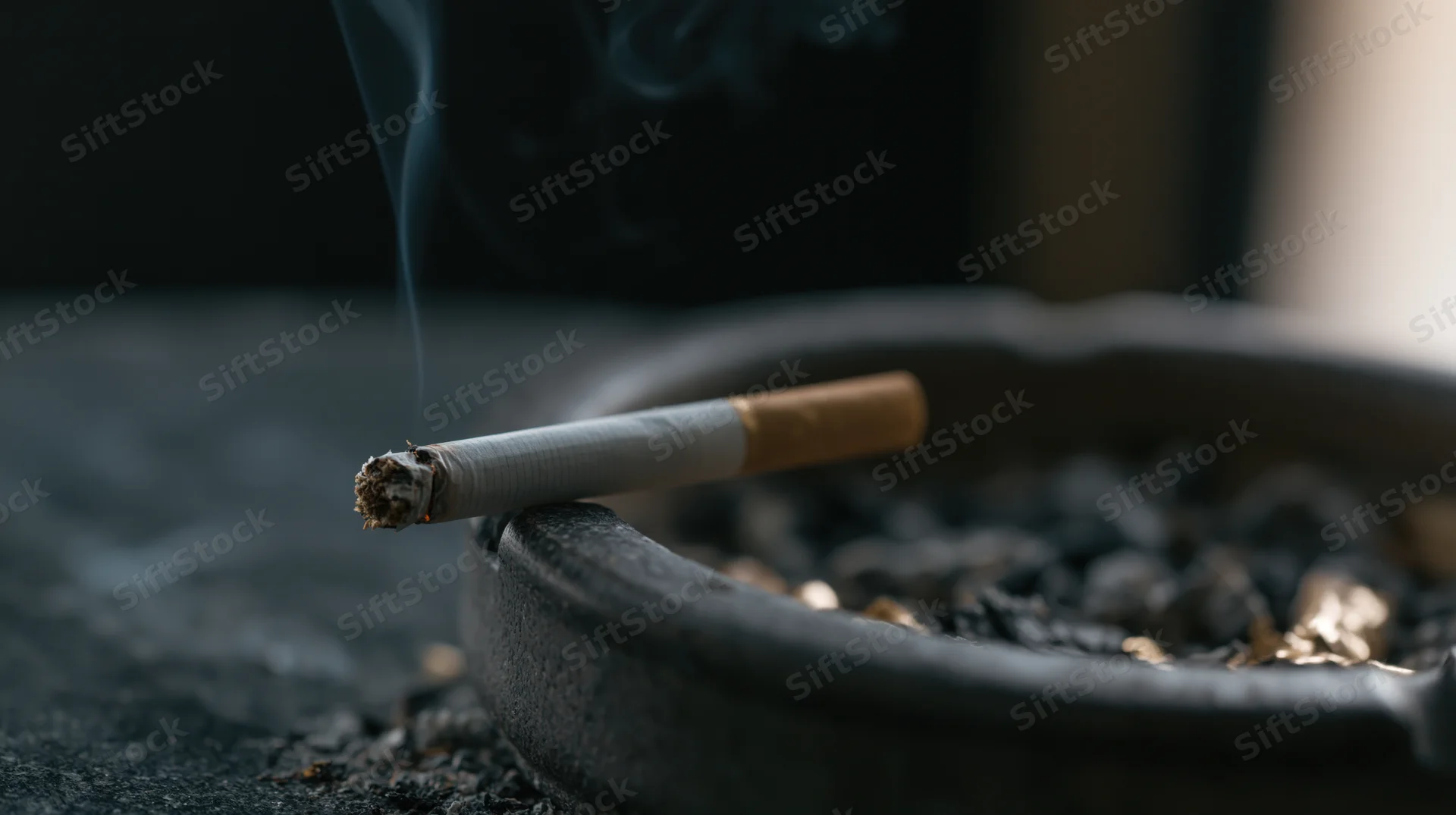 Lit cigarette resting on edge of dark ceramic ashtray with smoke rising