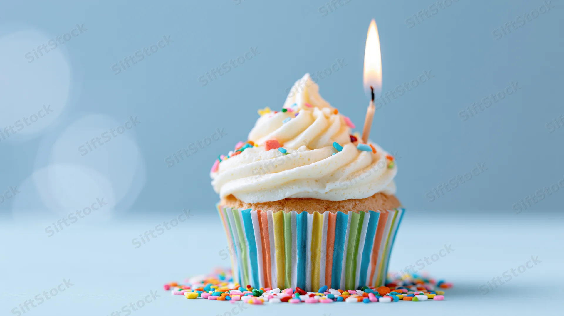 Single birthday cupcake with lit candle and sprinkles in rainbow striped paper liner on light blue background
