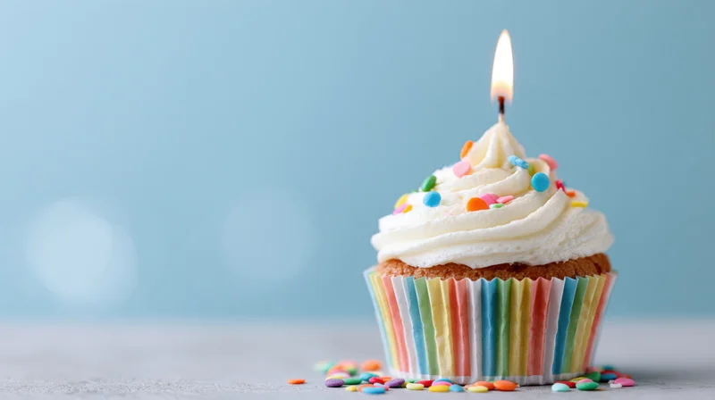 Single birthday cupcake with lit candle and sprinkles in rainbow striped paper liner on light blue background