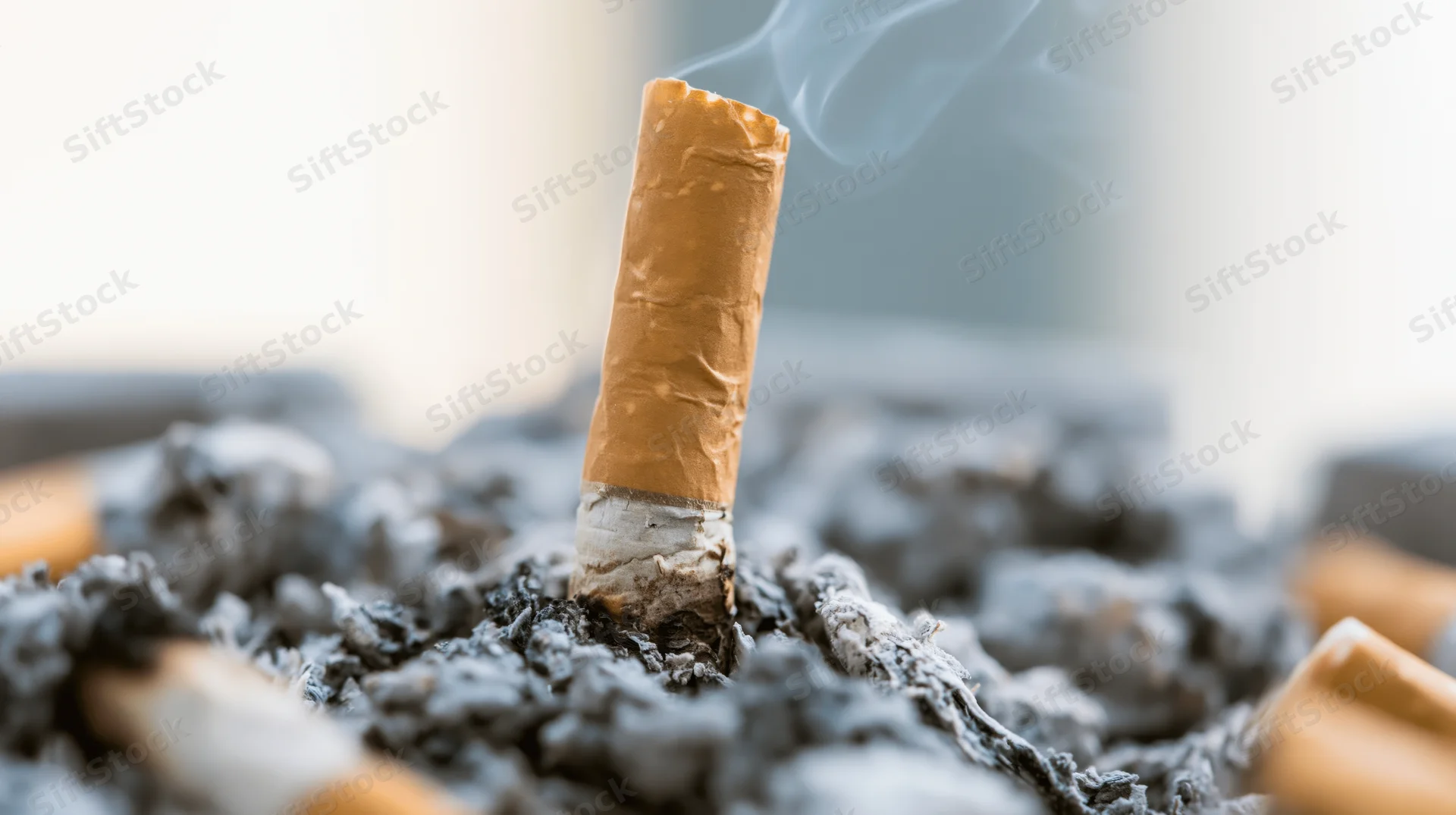Cigarette butt standing upright in ash with smoke rising