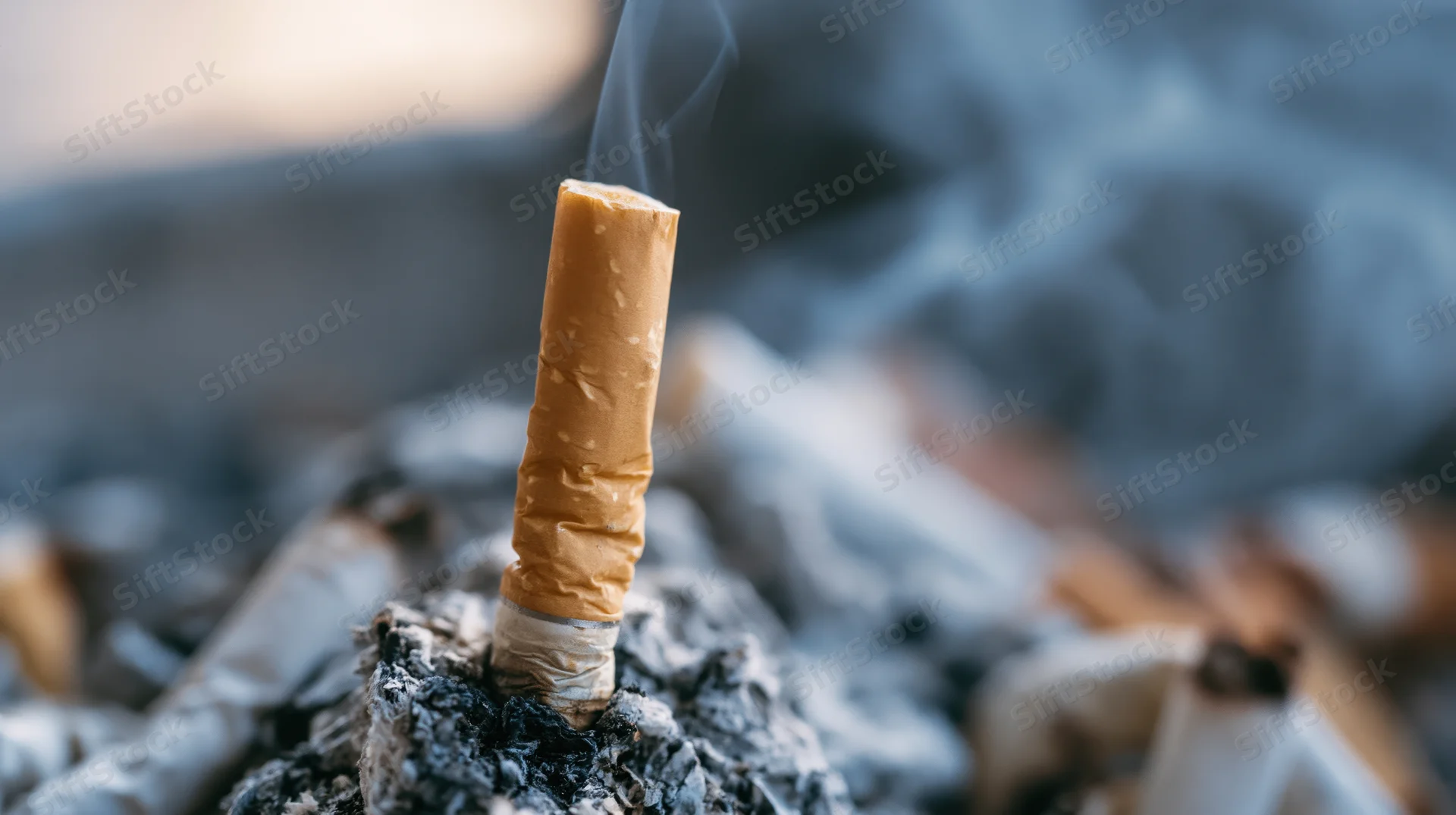 Cigarette butt standing upright in ash with smoke rising