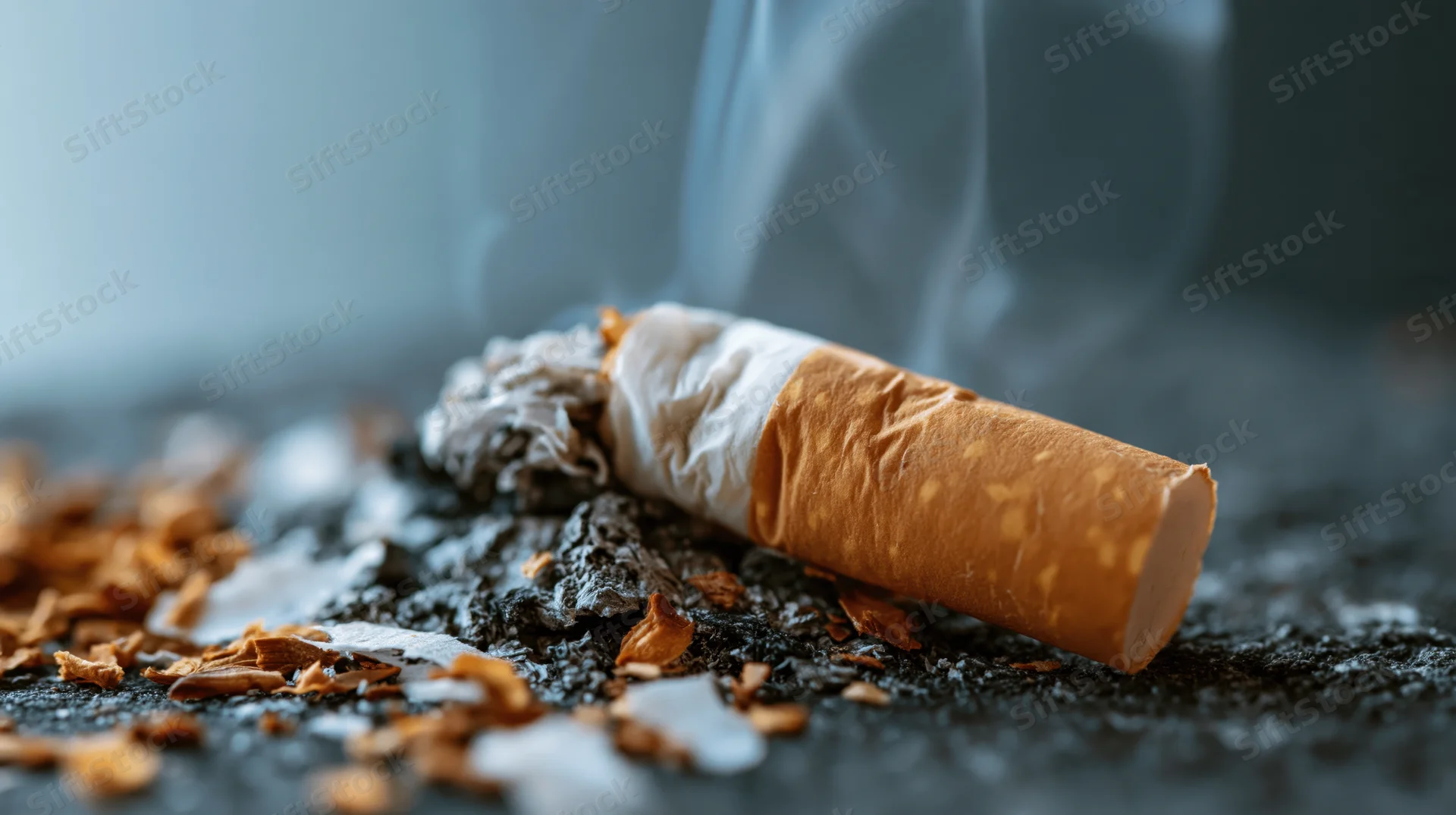 Crushed cigarette stub lying on dark ash-covered surface with smoke rising