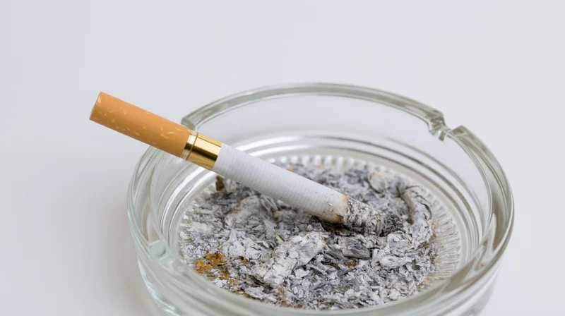Cigarette with gold filter resting in a glass ashtray filled with ash on a white background