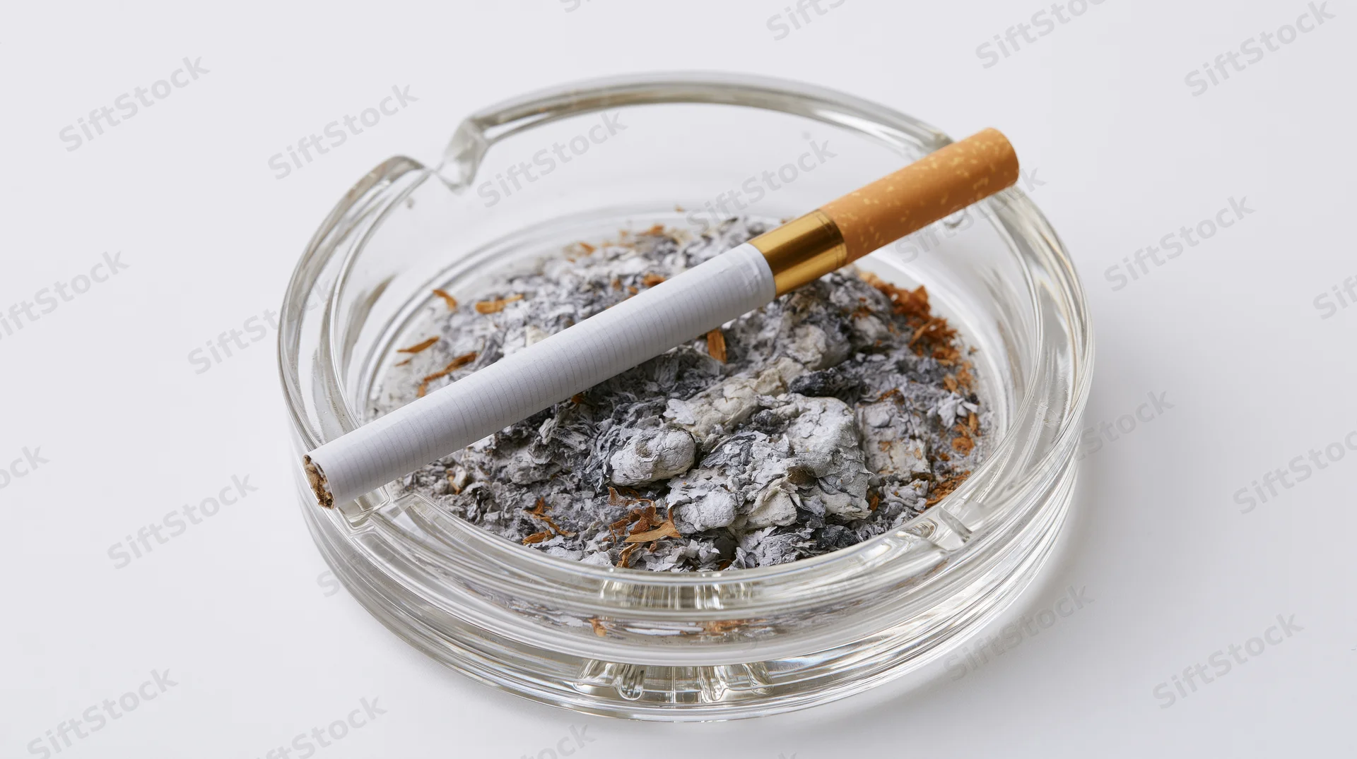 A cigarette with gold filter resting on a glass ashtray filled with ash and cigarette butts, isolated on white