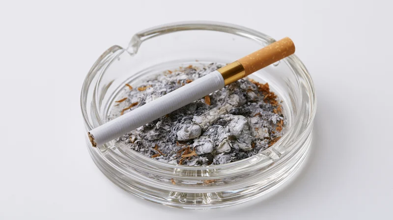 A cigarette with gold filter resting on a glass ashtray filled with ash and cigarette butts, isolated on white