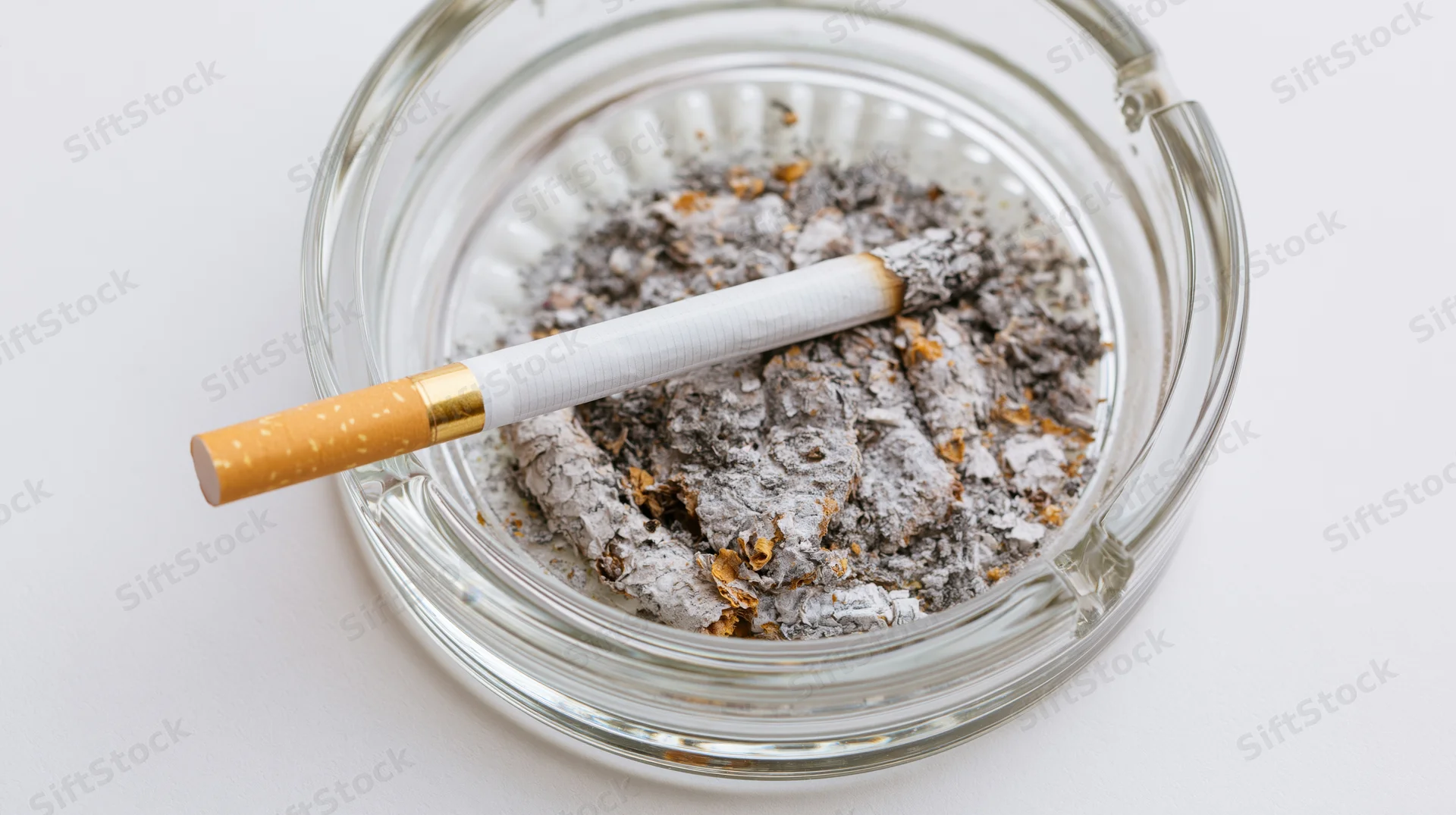 A cigarette with gold filter resting on a pile of ash in a clear glass ashtray on a white surface