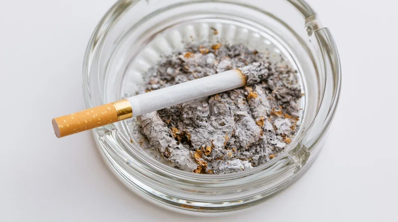 A cigarette with gold filter resting on a pile of ash in a clear glass ashtray on a white surface