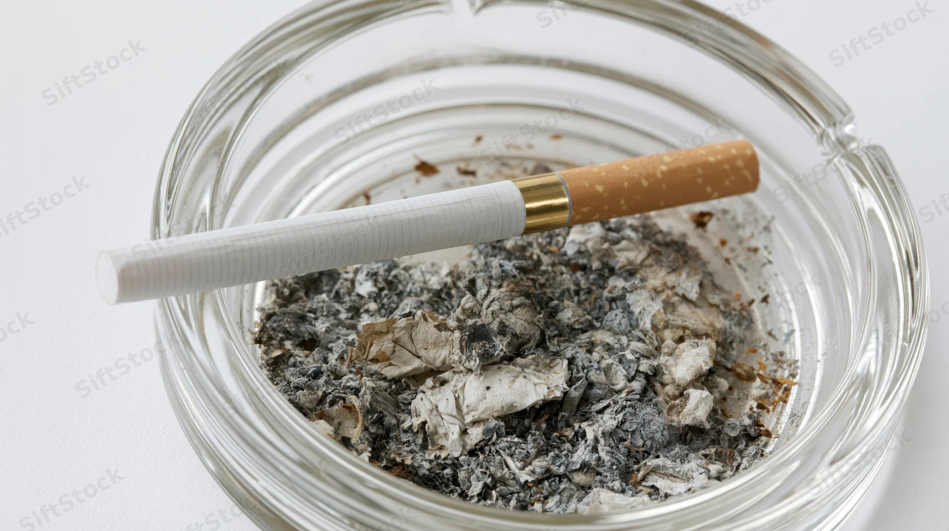 Cigarette with gold filter resting across a glass ashtray filled with ash on a white surface