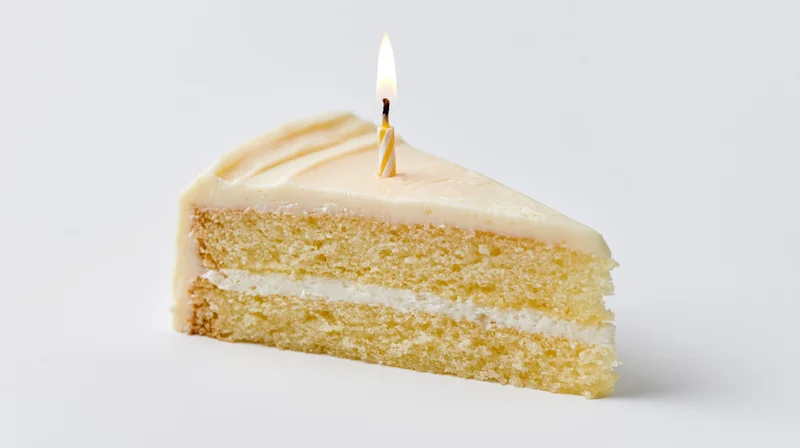 Single slice of triple-layer vanilla cake with lit candle on white background