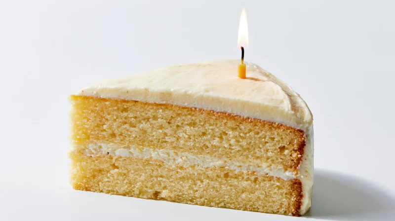 Triangular slice of triple-layer vanilla cake with lit candle on white background