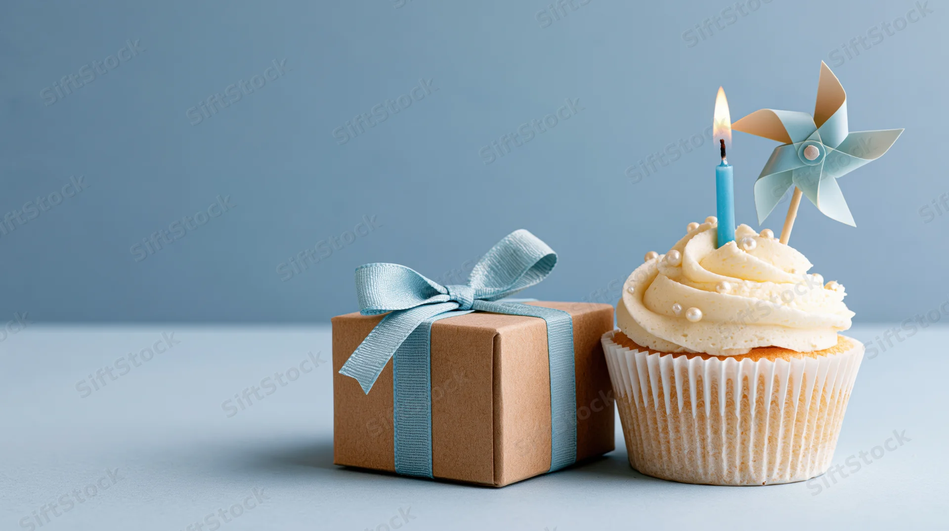 Cupcake with lit candle and pinwheel next to a kraft gift box tied with blue ribbon on a light blue surface