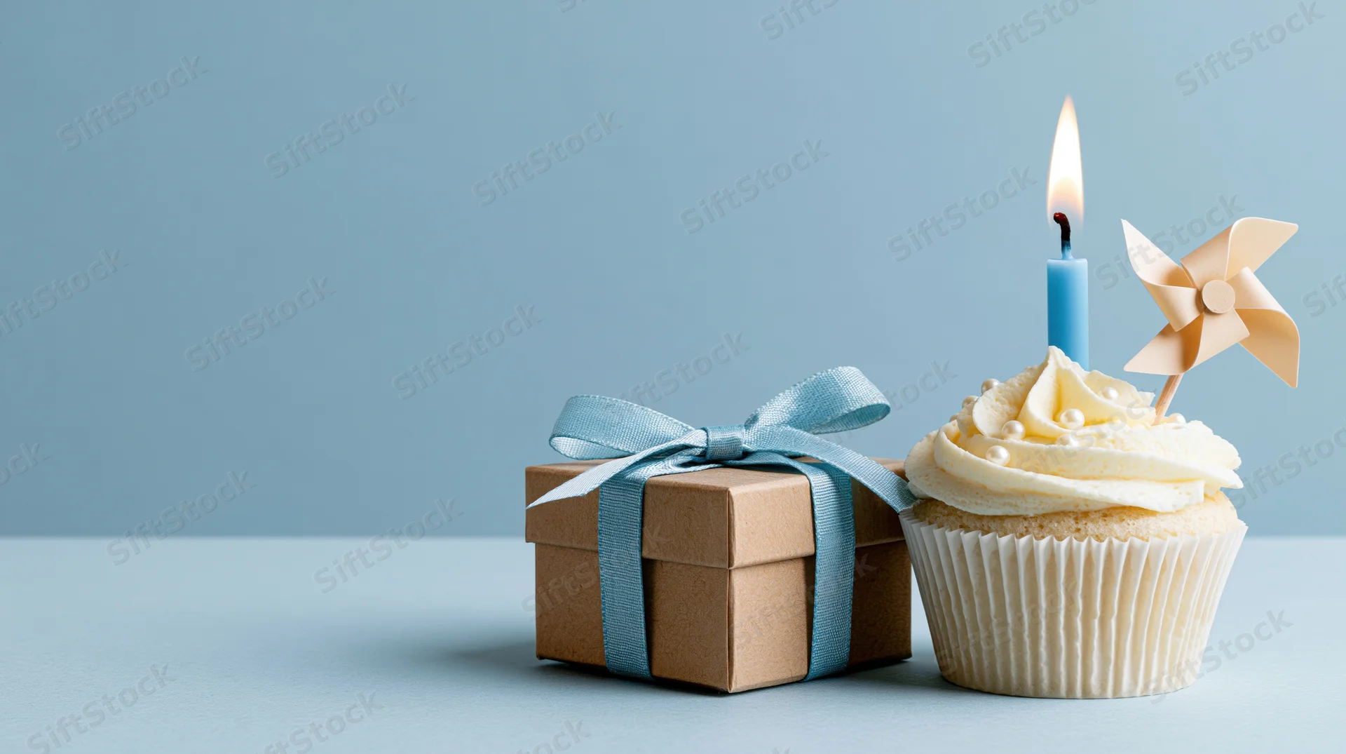 Cupcake with lit candle and pinwheel next to a kraft gift box tied with blue ribbon on a light blue surface