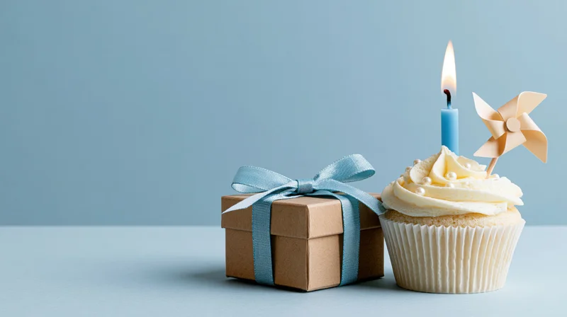 Cupcake with lit candle and pinwheel next to a kraft gift box tied with blue ribbon on a light blue surface