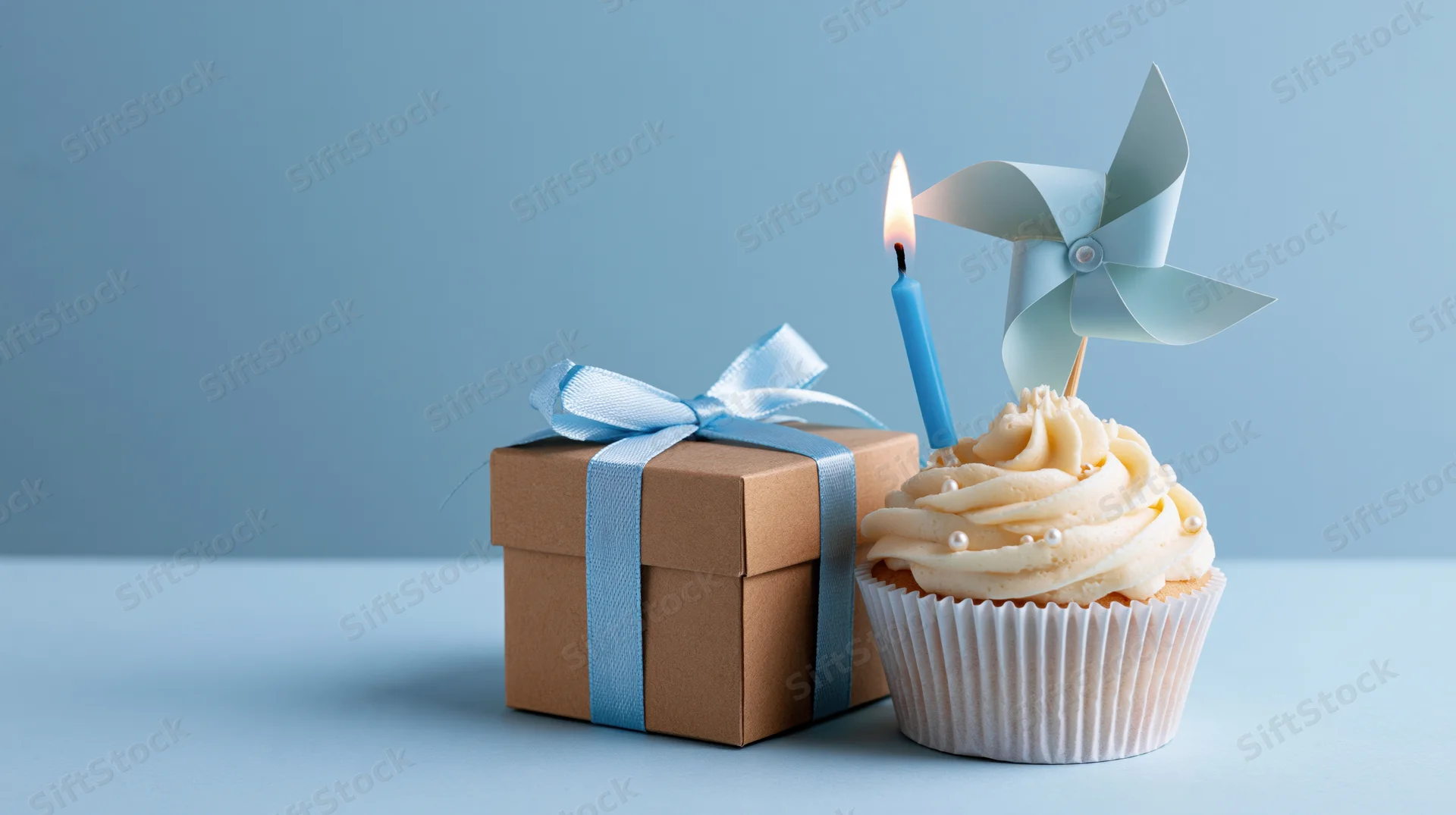 Cupcake with lit candle and pinwheel next to a kraft gift box tied with blue ribbon on a blue surface