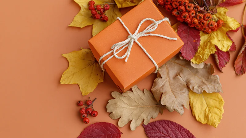 Orange gift box tied with white twine bow surrounded by autumn leaves and berries on warm-toned surface