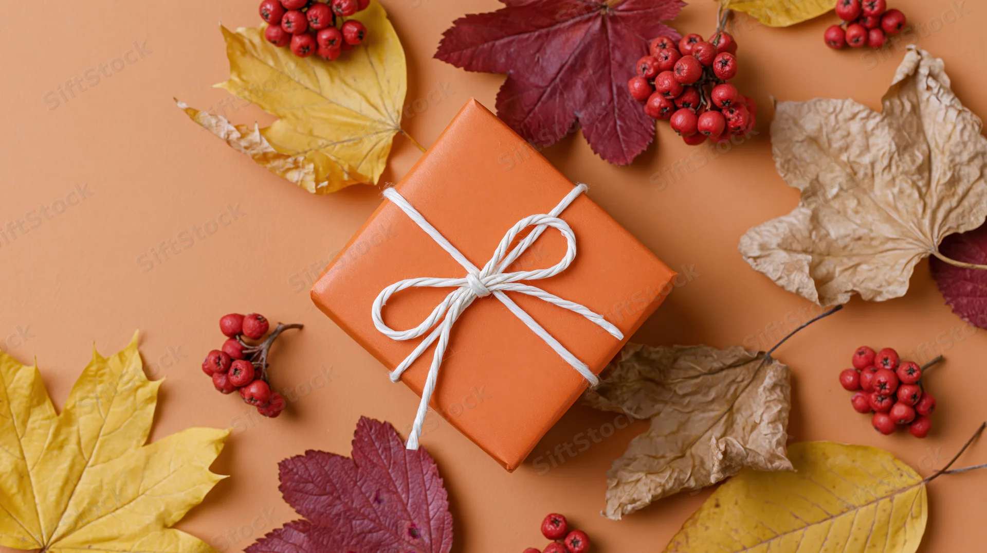 Orange gift box tied with white twine bow surrounded by autumn leaves and red berries on warm-toned surface