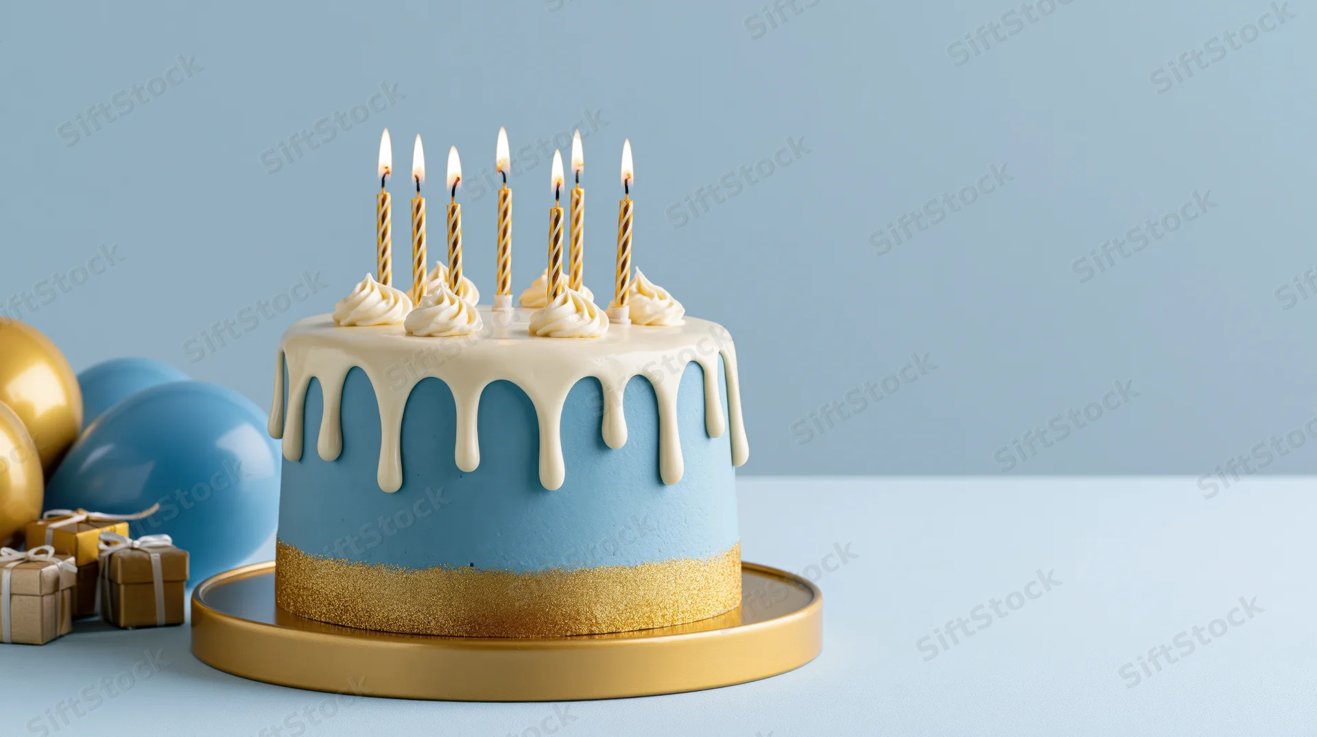 Blue and gold birthday cake with lit candles on a plate, surrounded by balloons and gifts on a light blue surface