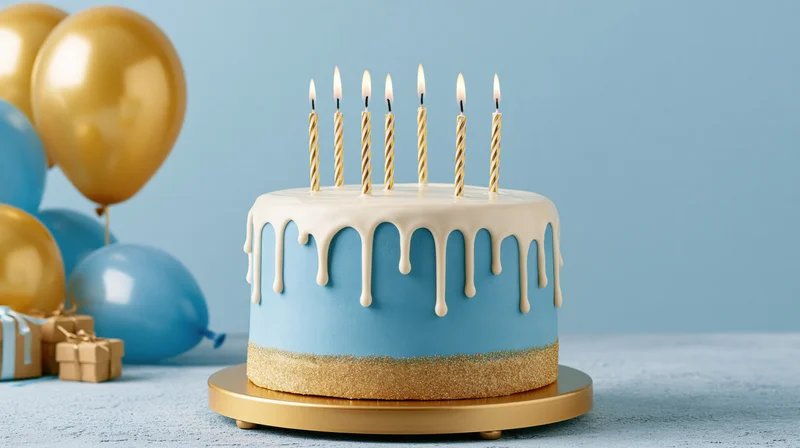 Blue and gold birthday cake with lit candles on a stand, surrounded by balloons and gifts on a table