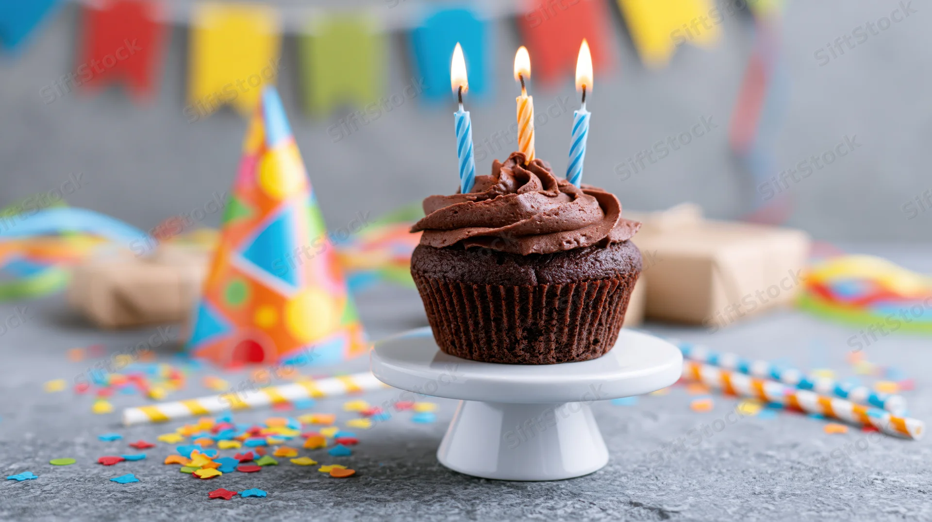 Chocolate cupcake with lit candles on a white stand surrounded by party decorations