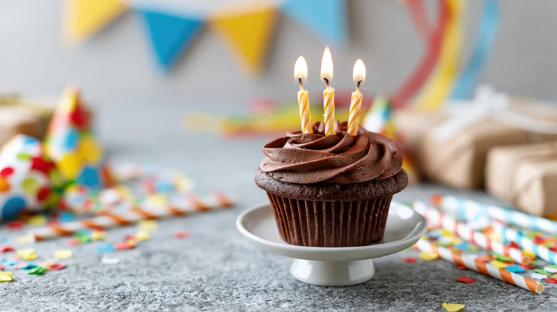 Chocolate cupcake with lit twisted candles on a small white stand surrounded by party decorations on a gray surface