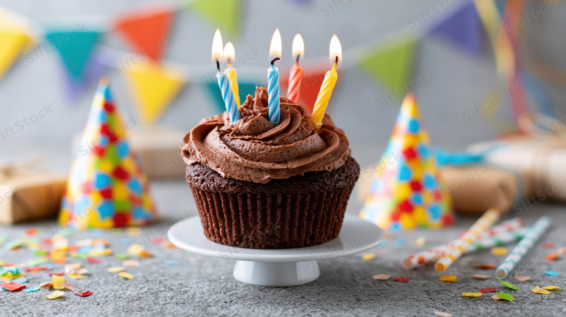 Chocolate cupcake with lit twisted birthday candles on a small white stand surrounded by party hats, confetti, and bunting flags
