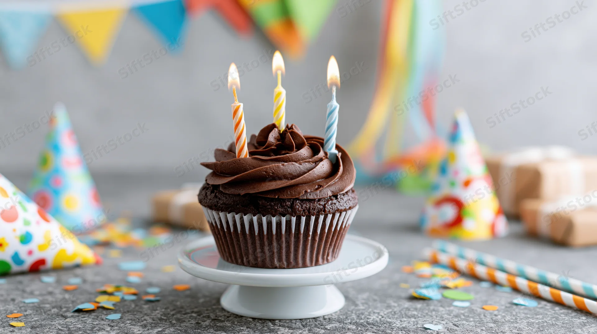 Chocolate cupcake with lit twisted birthday candles on a small white stand surrounded by party hats and confetti