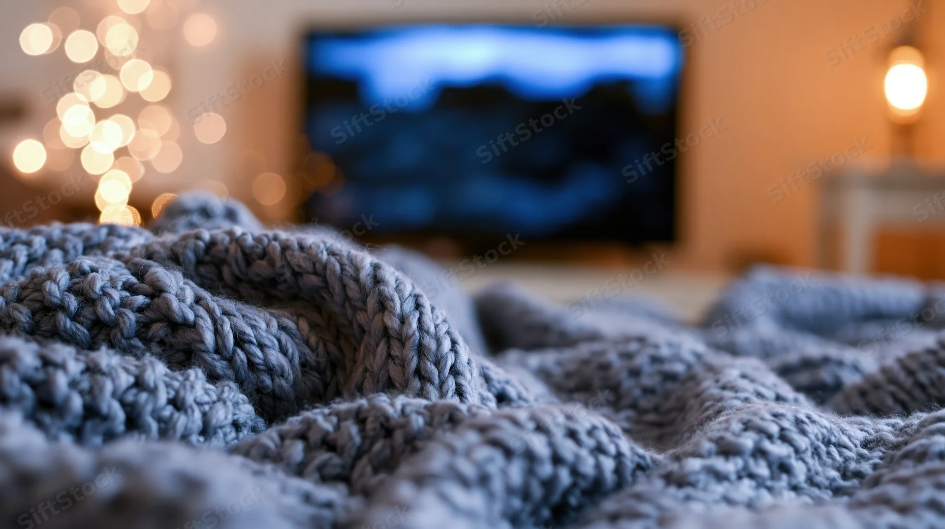 Close-up of a rumpled gray knit blanket in a living room with a television and festive lights in the background