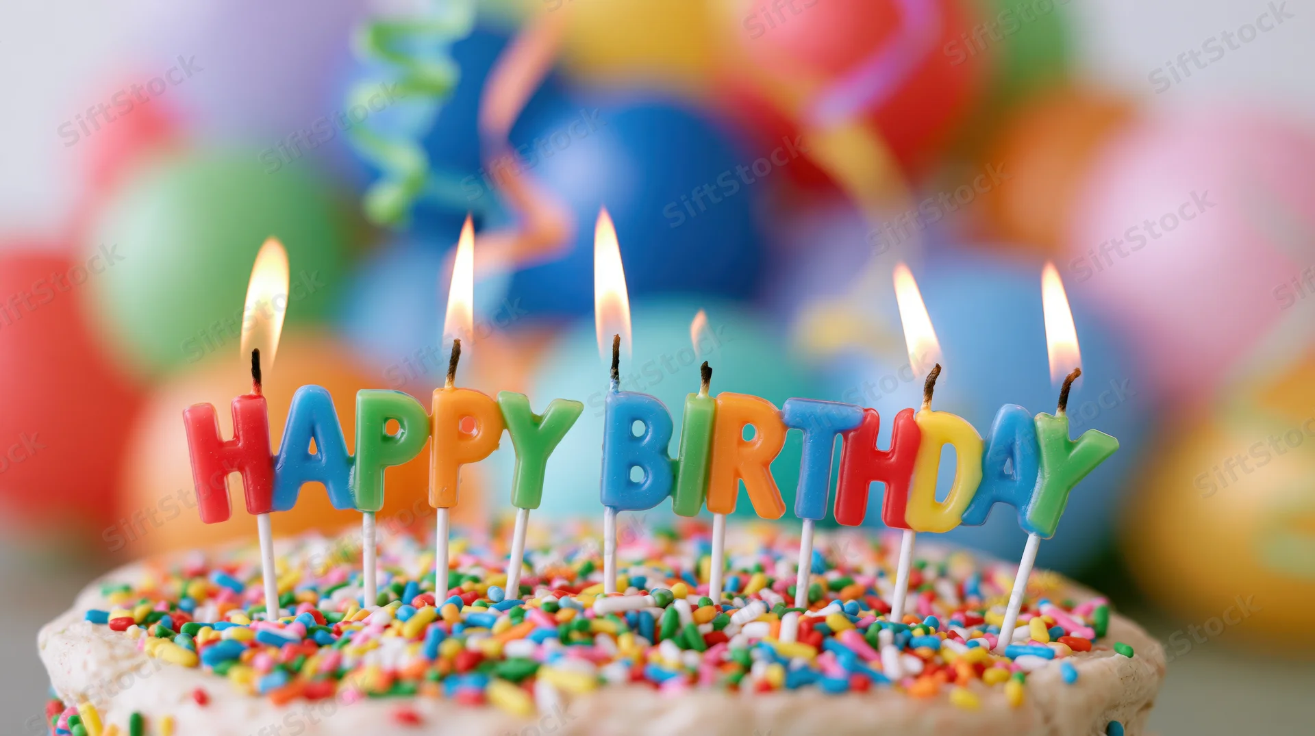Birthday cake with lit letter candles spelling happy birthday, decorated with colorful sprinkles, balloons and streamers in background