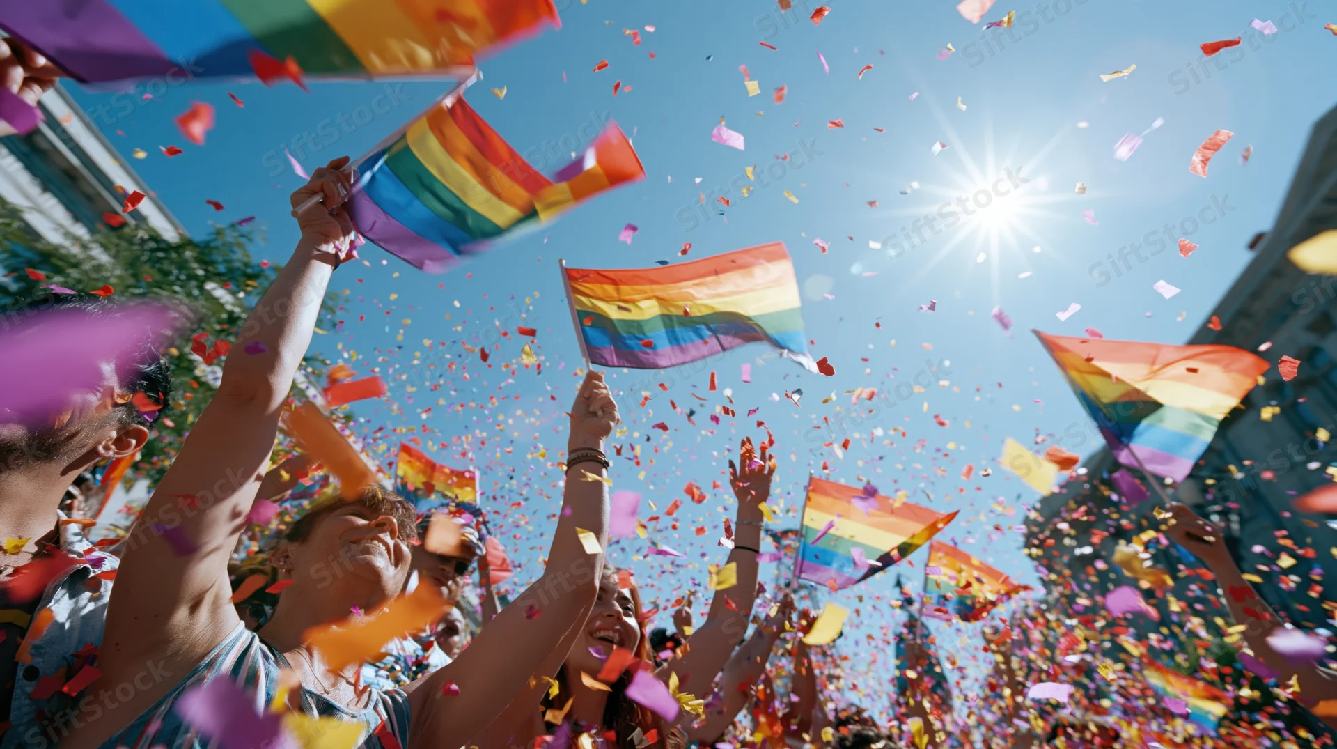 People waving rainbow flags amidst falling confetti at outdoor pride parade under bright sun