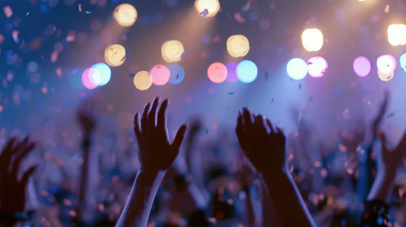 Silhouetted hands raised in a crowd at a concert with colorful bokeh lights and falling confetti