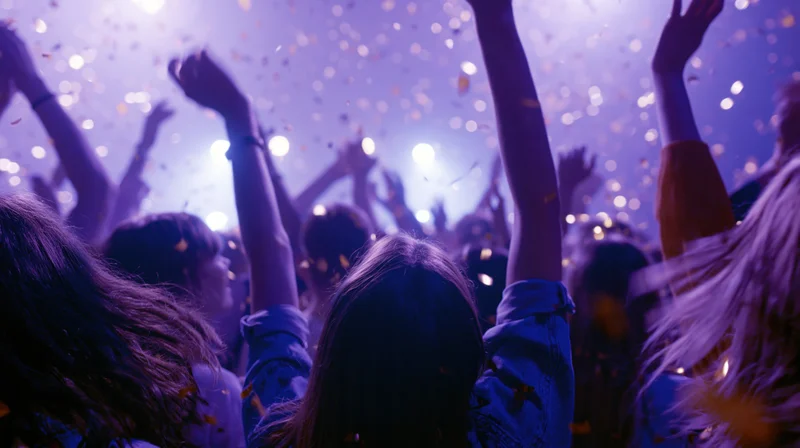 Crowd of young people dancing with arms raised at indoor nightclub concert under purple lights and falling confetti