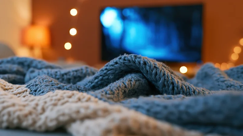 Crumpled blue and beige knit blanket in foreground with blurred television and warm lights in background