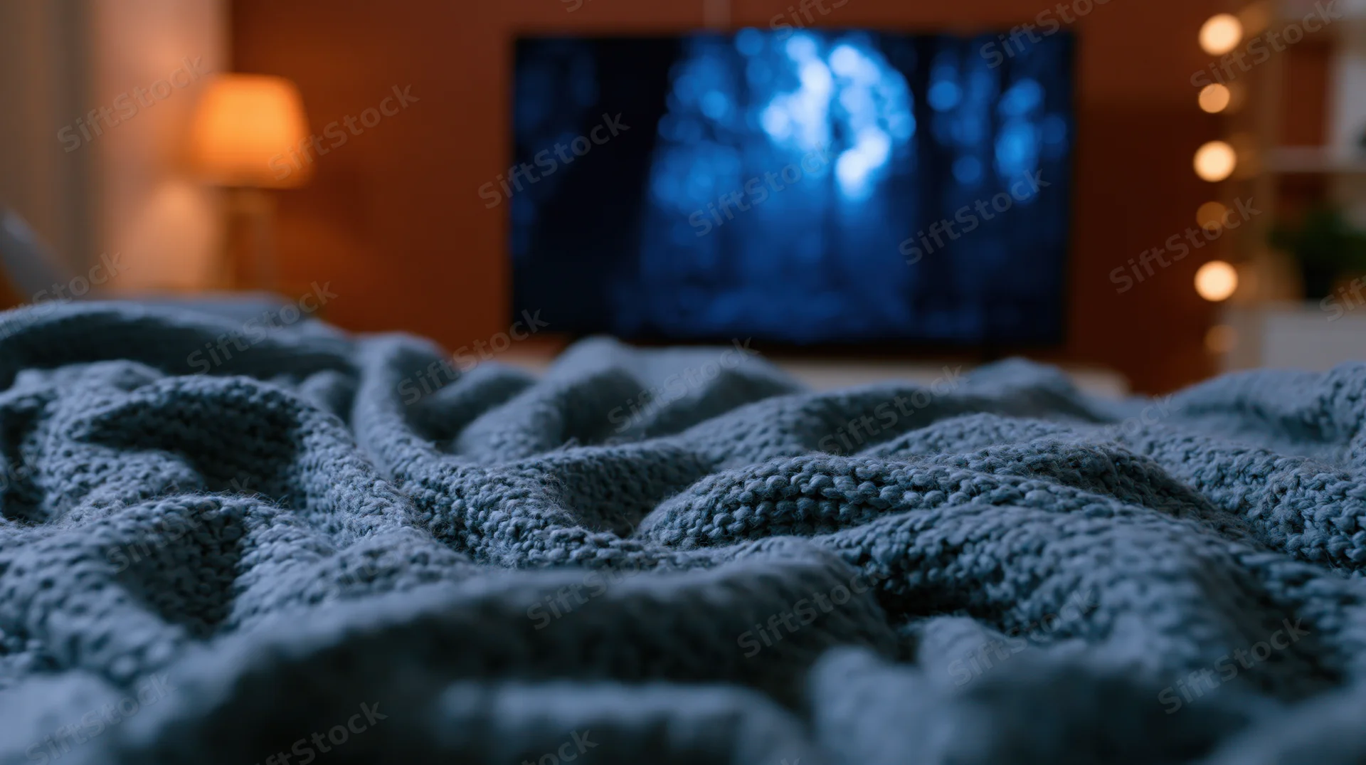 Crumpled blue knit blanket in foreground with television showing forest scene in dimly lit living room