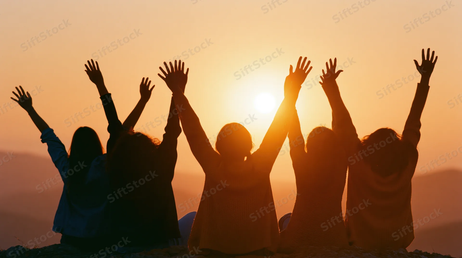 Five friends sitting on a hilltop with arms raised at sunset