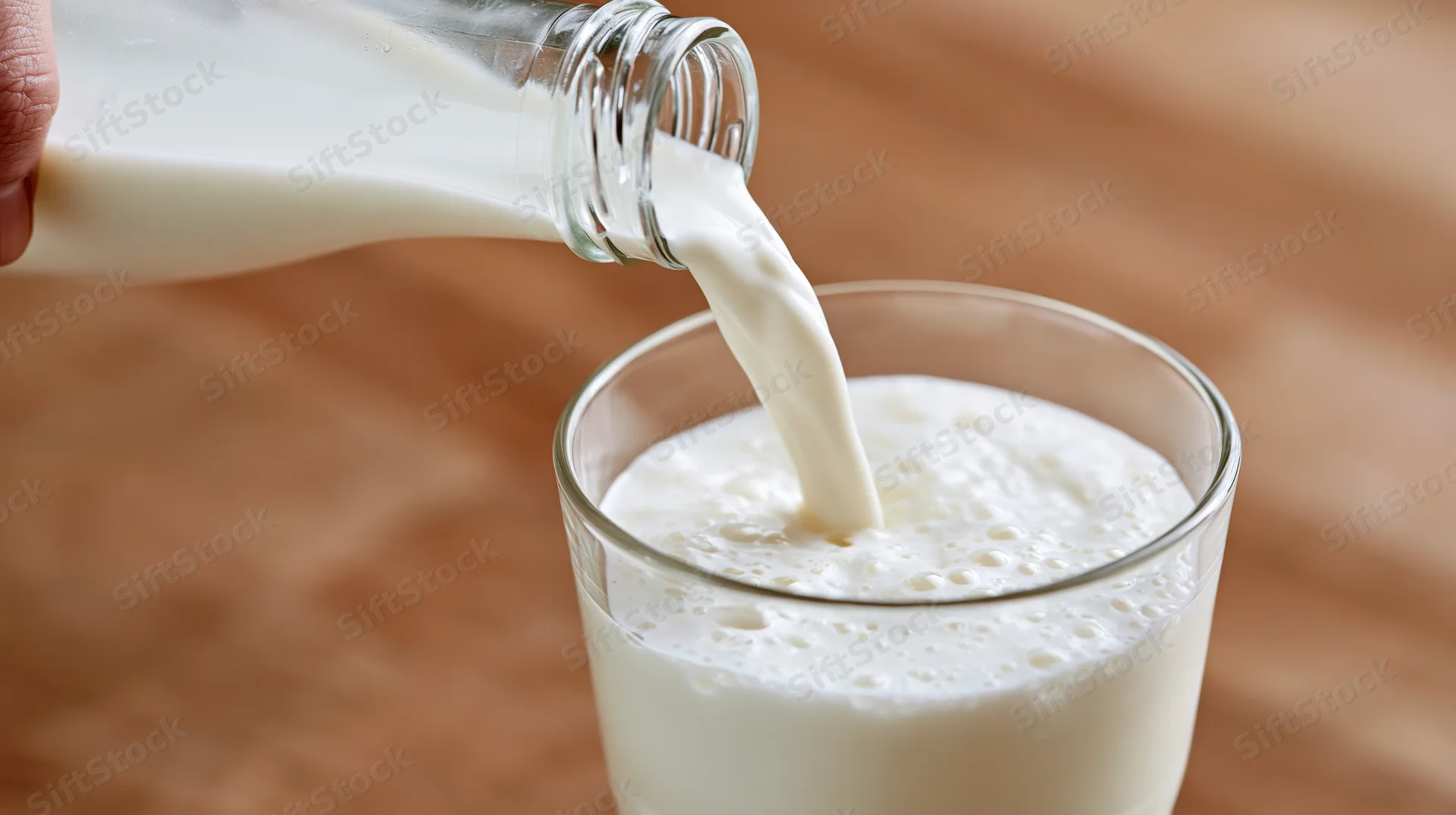 Hand pouring milk from a glass bottle into a clear glass on a wooden surface