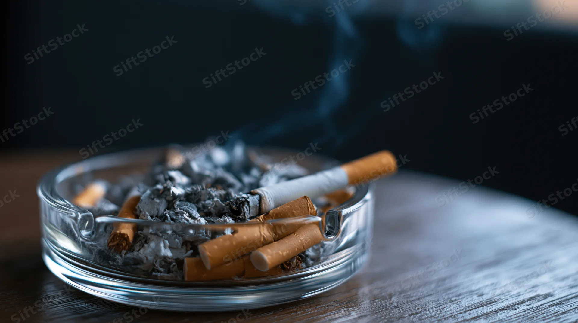 Glass ashtray filled with cigarette butts and ash on a wooden table with smoke rising