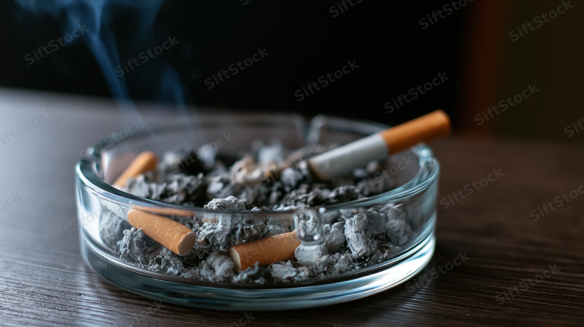Glass ashtray filled with cigarette butts and ash on wooden table with smoke rising