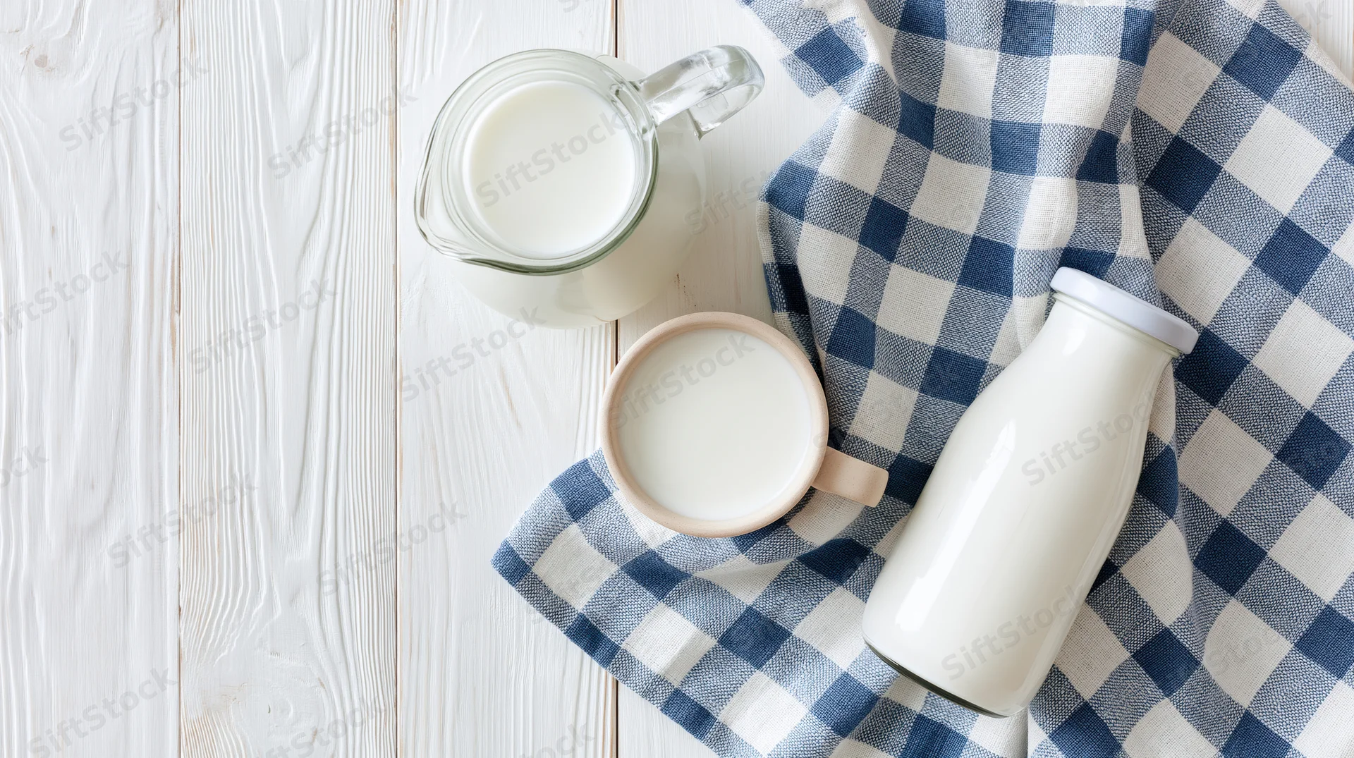 Milk in glass pitcher ceramic mug and sealed bottle arranged on white wooden table with blue checkered cloth