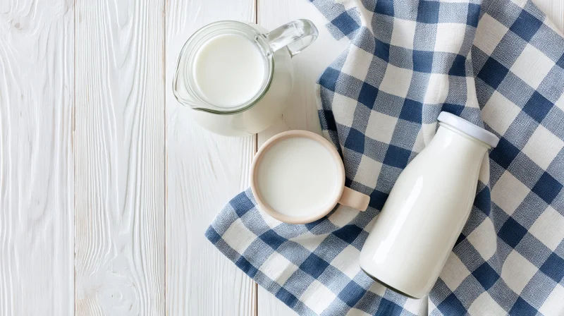 Milk in glass pitcher ceramic mug and sealed bottle arranged on white wooden table with blue checkered cloth