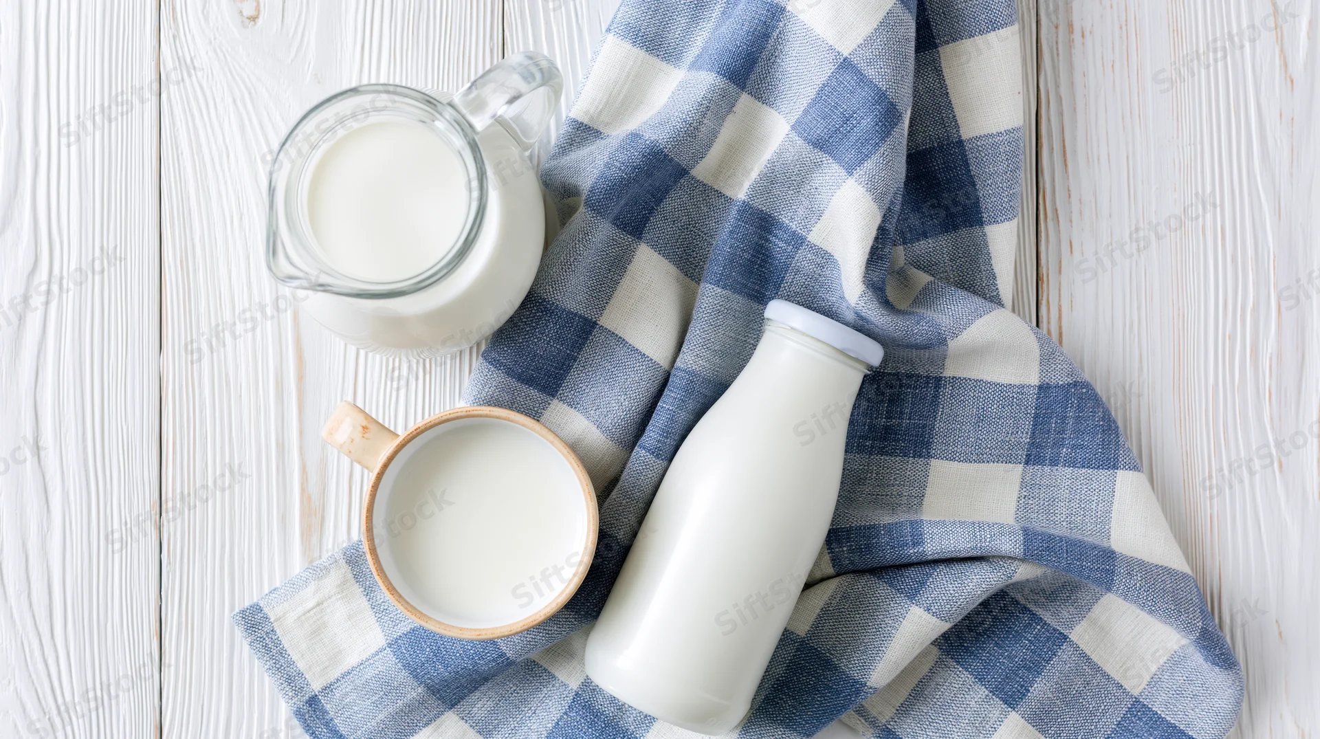 Milk in a glass pitcher, ceramic mug and sealed bottle arranged on a checkered cloth over a white wooden table
