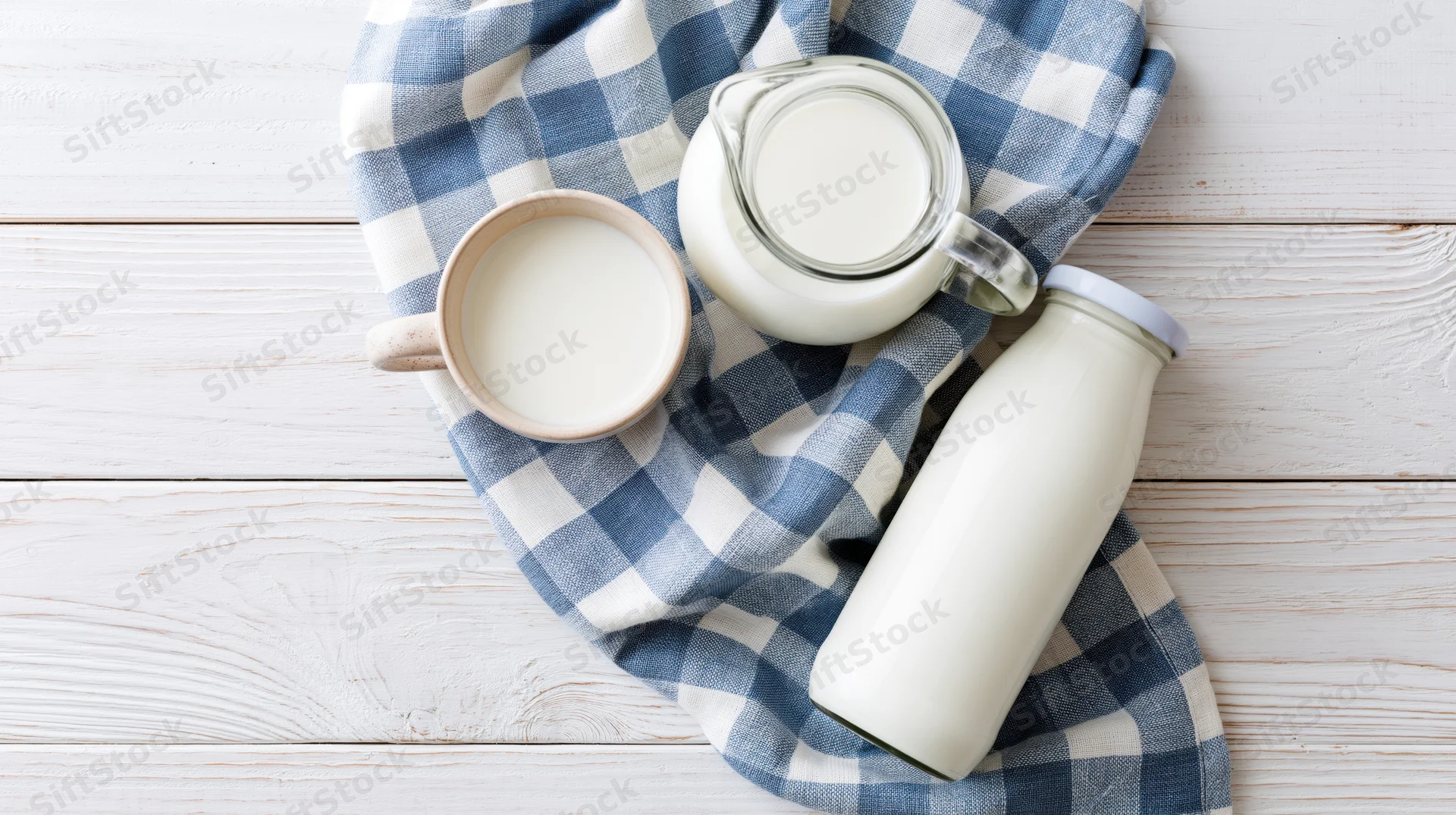 Milk in glass pitcher ceramic mug and sealed bottle on checkered cloth over white wooden table