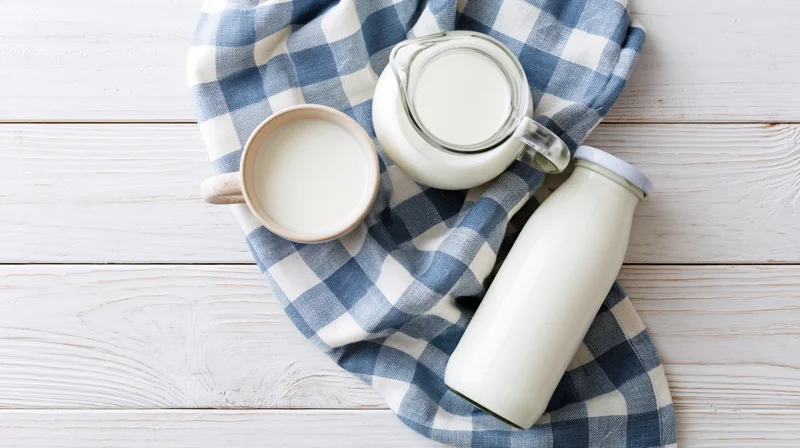 Milk in glass pitcher ceramic mug and sealed bottle on checkered cloth over white wooden table