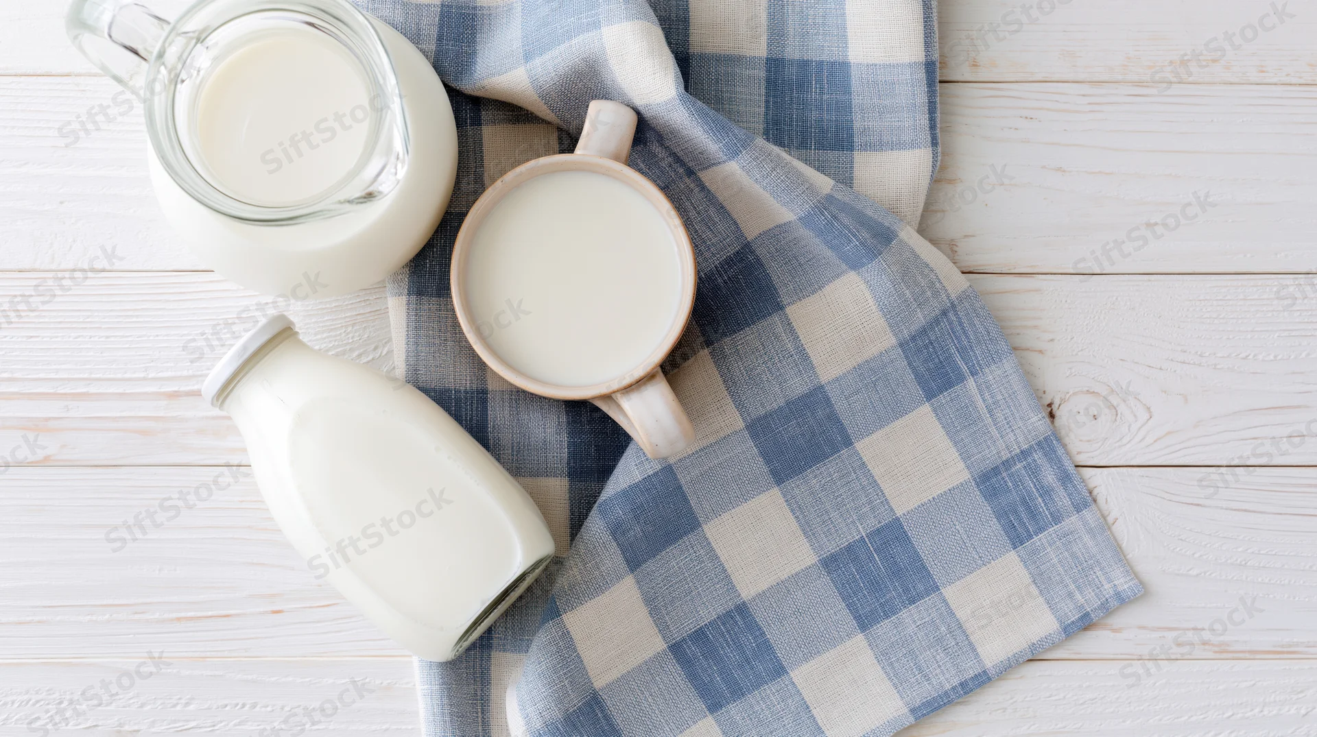 Milk in glass pitcher ceramic mug and sealed bottle arranged on blue checkered cloth over white wooden table