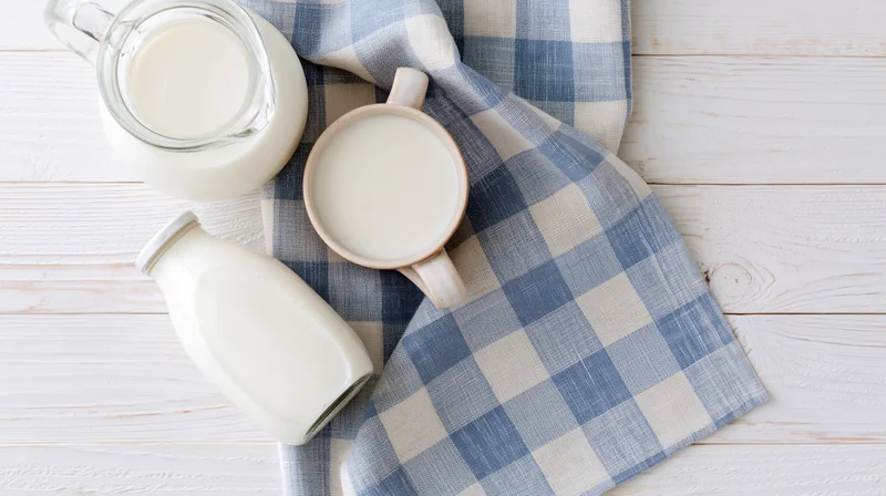 Milk in glass pitcher ceramic mug and sealed bottle arranged on blue checkered cloth over white wooden table
