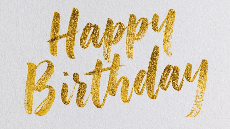 Hand-lettered happy birthday calligraphy in gold glitter on textured white paper