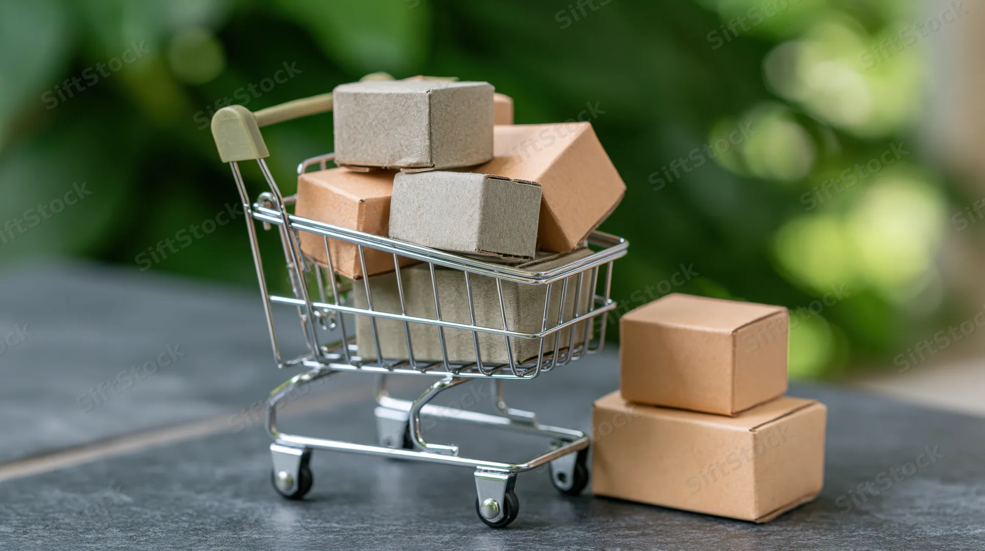 Miniature metal shopping cart filled with small cardboard boxes on a gray surface with green foliage in the background