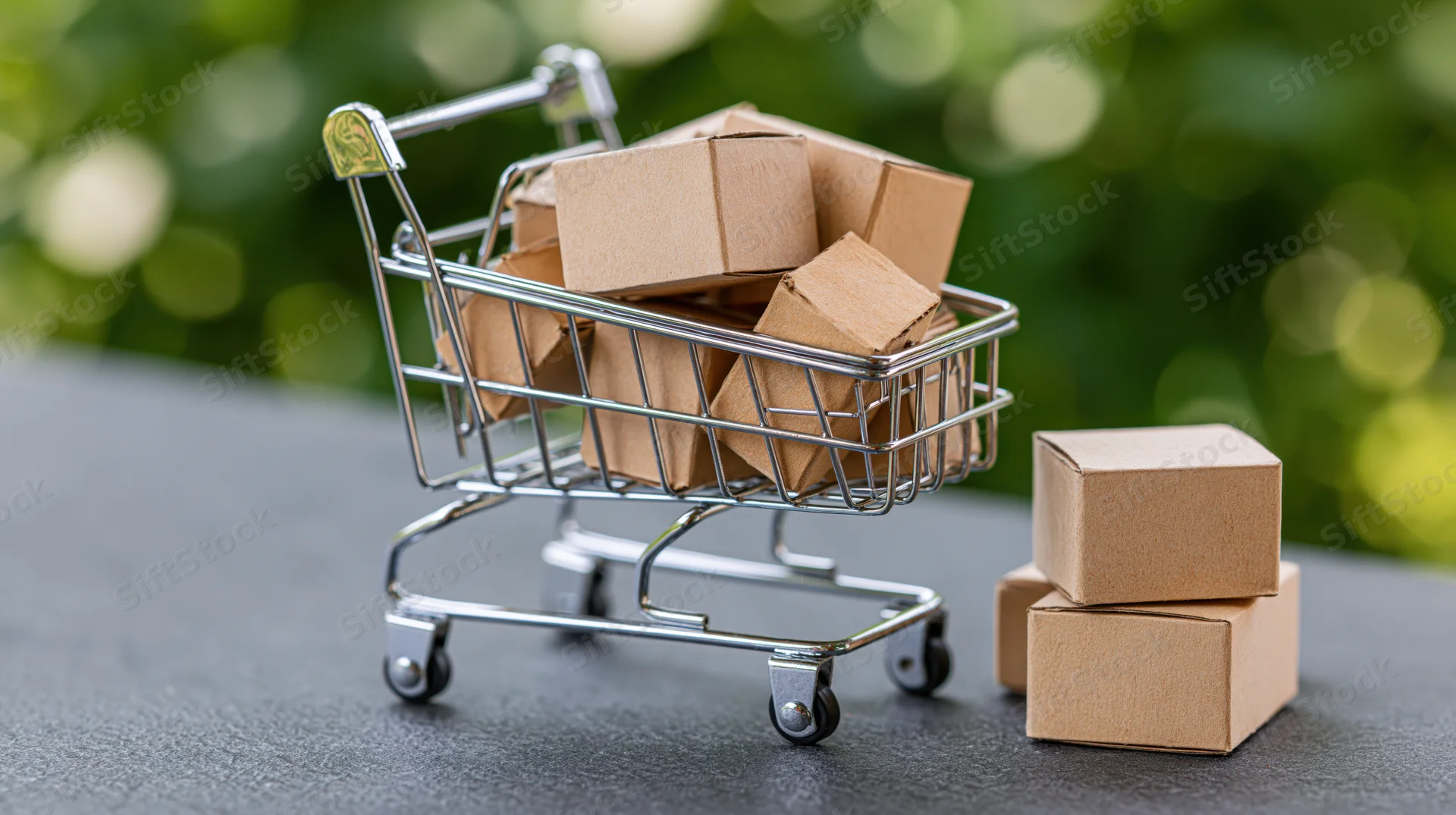 Miniature metal shopping cart filled with small cardboard boxes on a dark surface with blurred green background