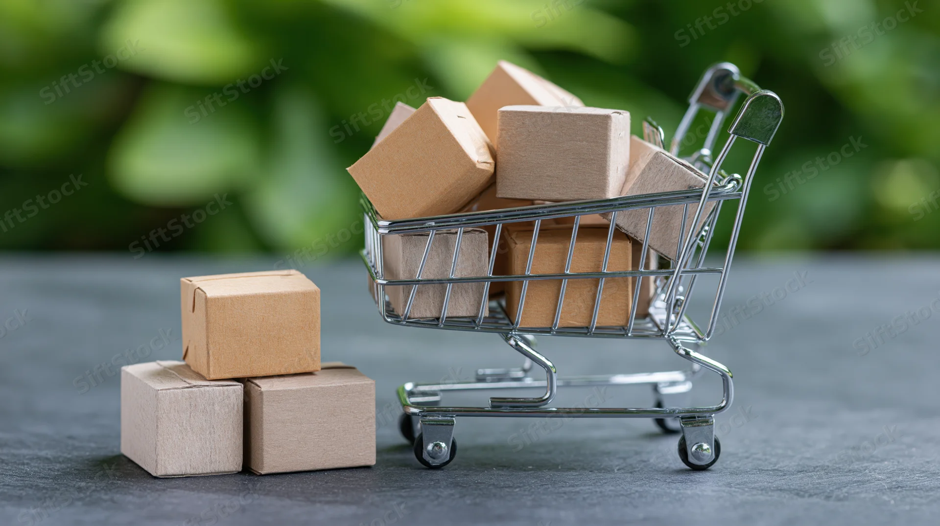 Miniature metal shopping cart filled with small cardboard boxes on a gray surface with green foliage background