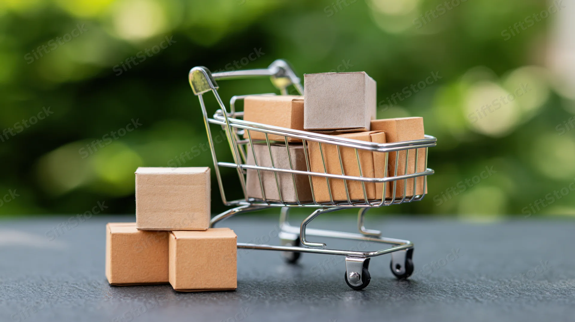 Miniature metal shopping cart filled with small cardboard boxes on dark surface with green blurred background